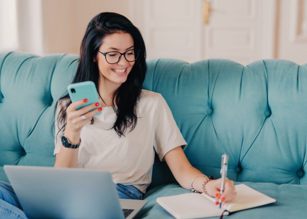 Smiling woman sitting on a teal couch using her phone while writing in a notebook with a laptop in front of her working on digital marketing or content creation strategy.